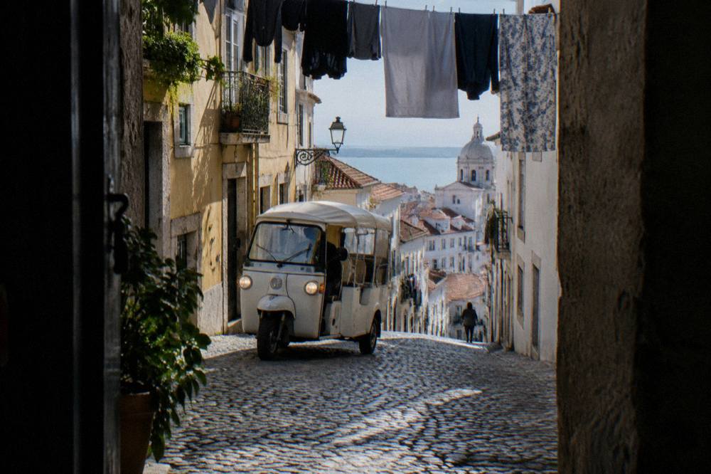 An electric tuk tuk driving up a narrow cobbled street between old tiled buildings in Lisbon