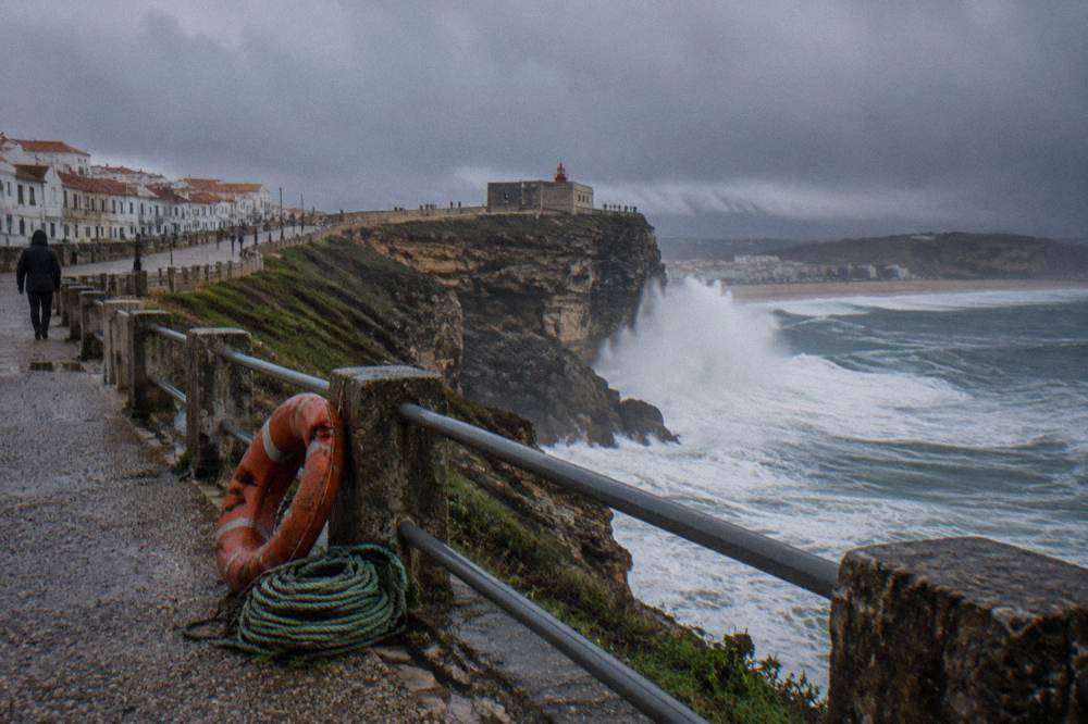 The giant waves of Nazare crashing against the cliffs with the red lighthouse in the distance