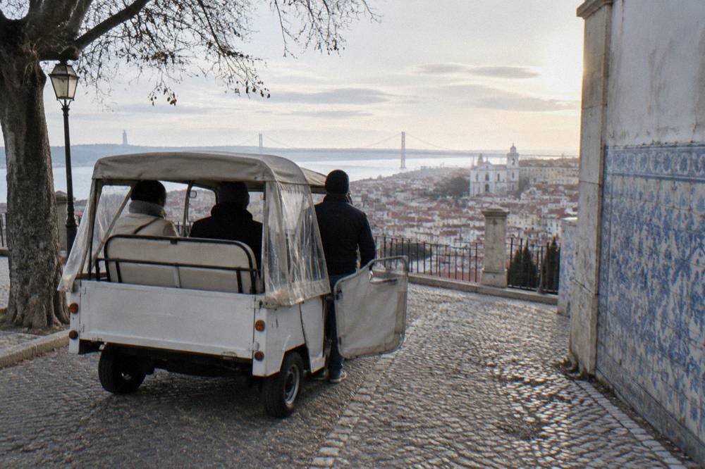 A tuk tuk driver speaking with passengers beside a tiled building on a Lisbon hillside street