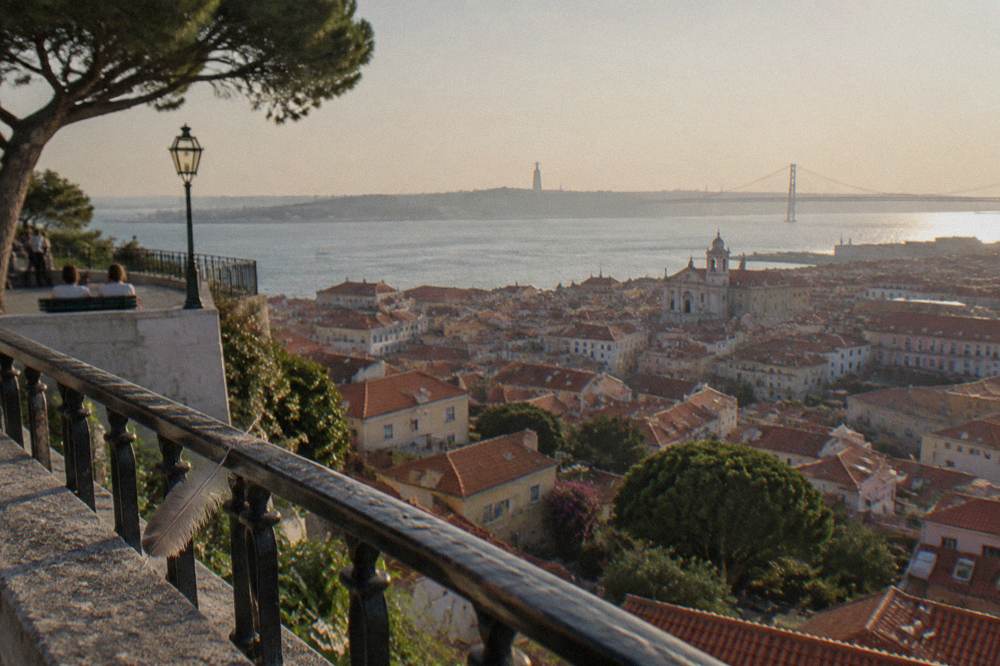 A panoramic view over Lisbon's red-tiled rooftops from a high miradouro, with the Tagus River in the distance under a blue sky