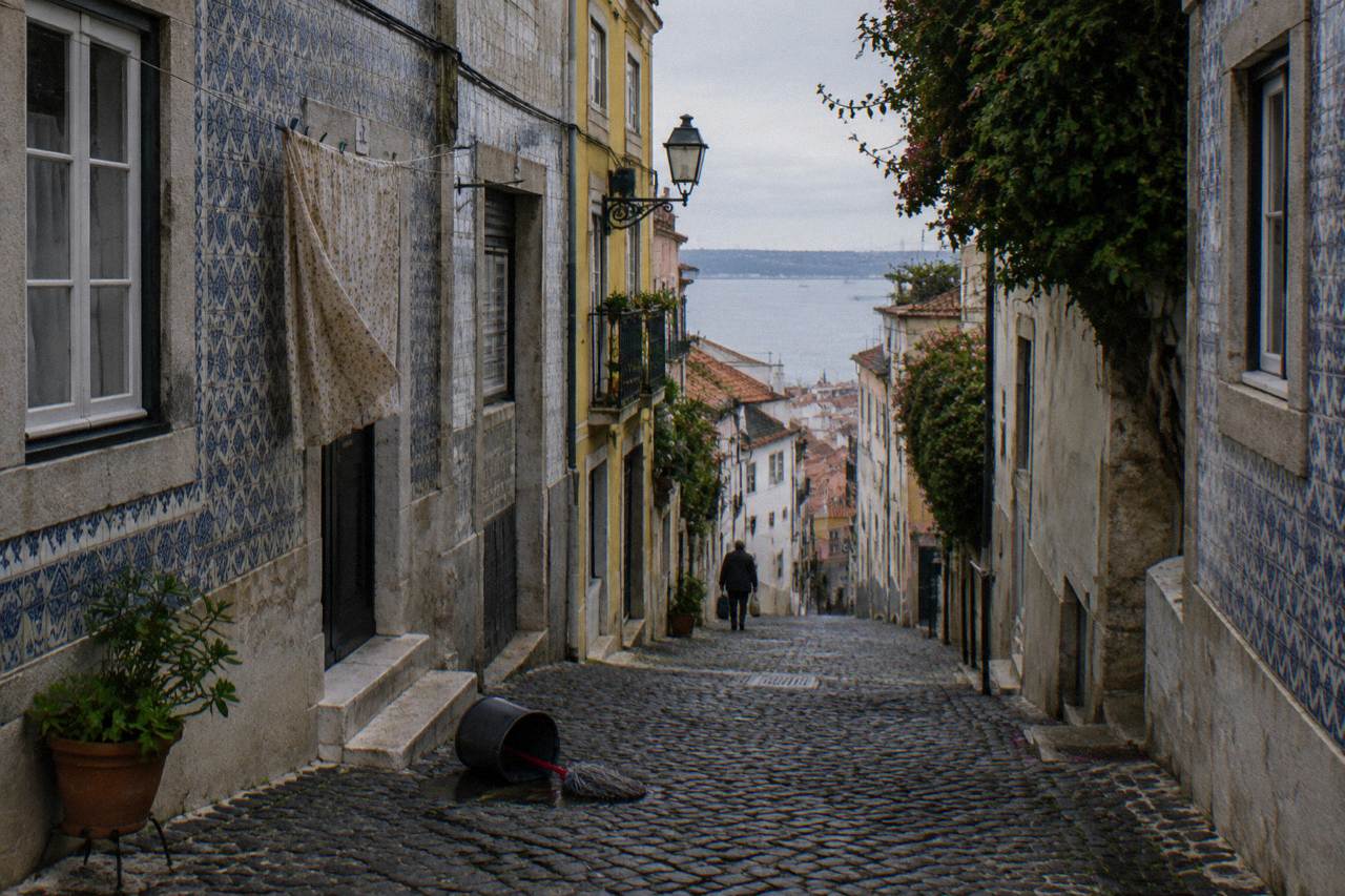 Narrow cobbled street in old Lisbon with tiled building facades and a distant river view