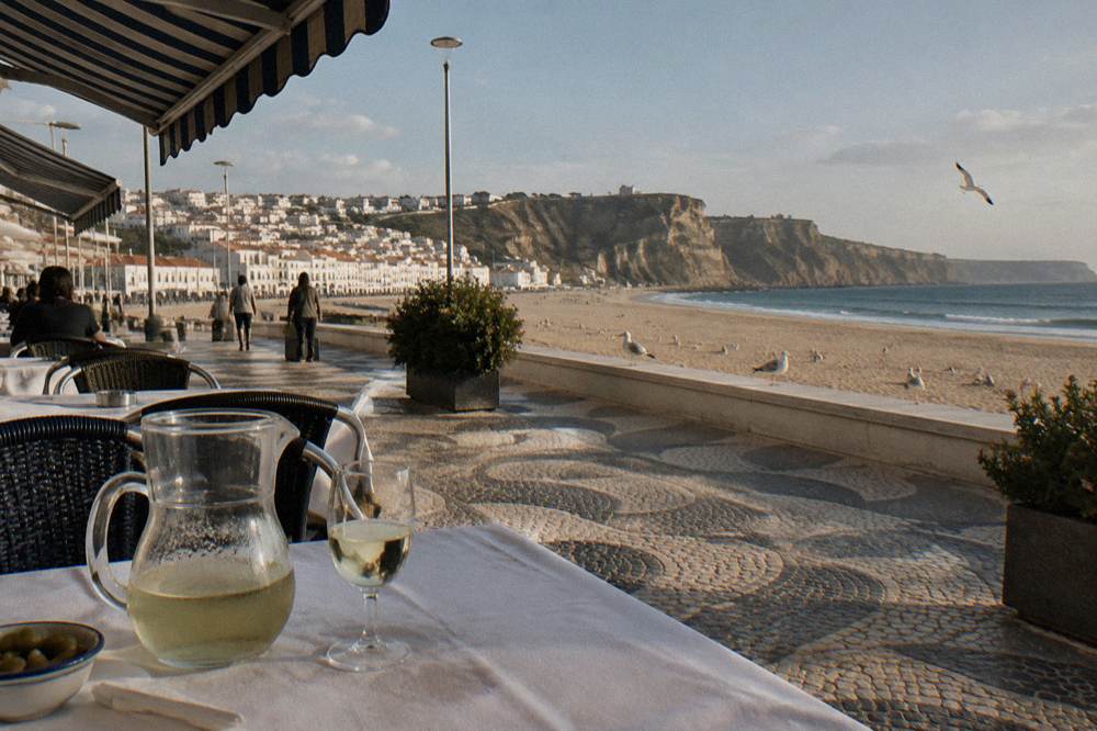 Seafront promenade and restaurant terraces along the wide beach at Nazare on a bright calm afternoon.