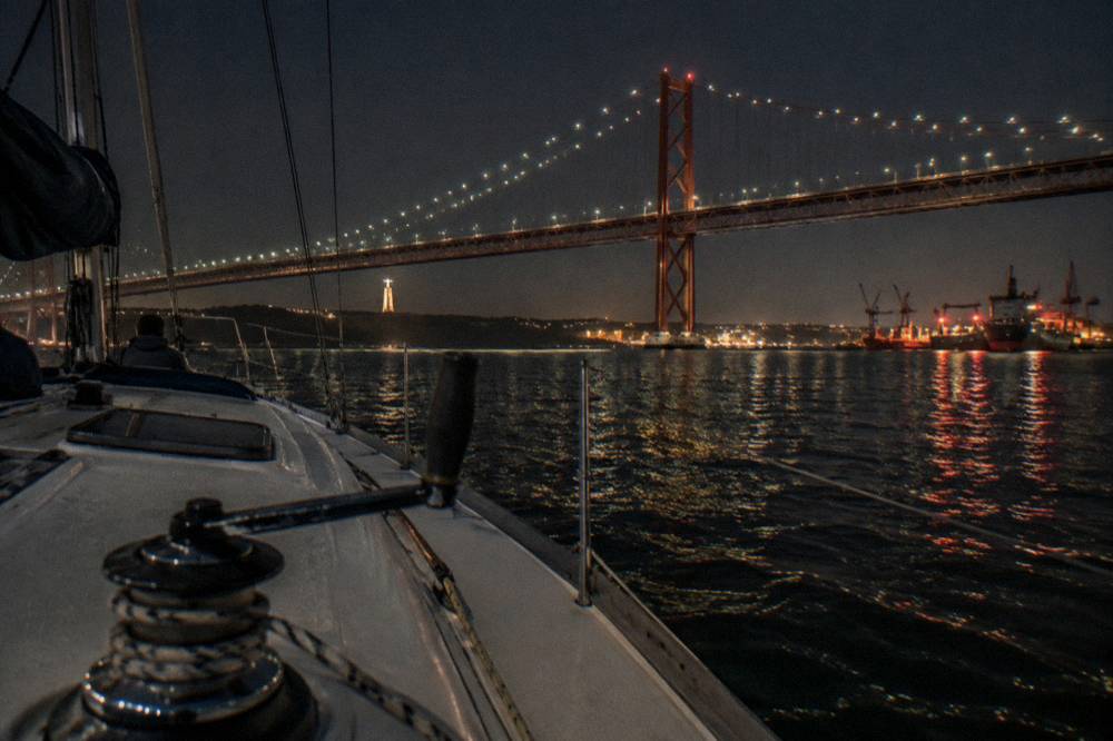 A sailing yacht passing under the illuminated 25 de Abril bridge at dusk