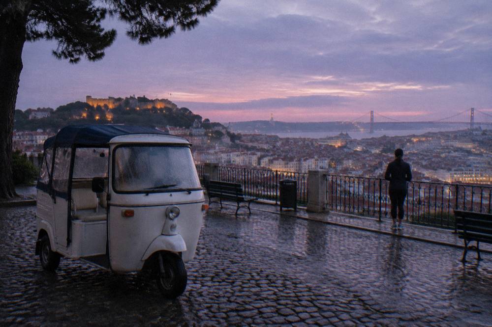A three-wheeled electric tuk tuk parked on a cobbled viewpoint above old Lisbon rooftops